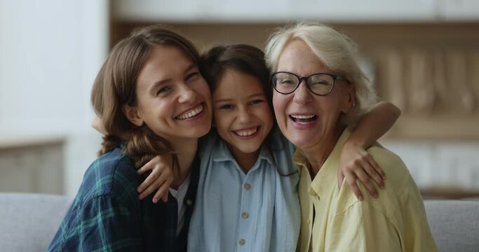 Close up portrait three loving female generations look at camera, pose for picture, enjoy leisure and priceless time together, cute girl hug mom and granny in living room shows family unity and bond