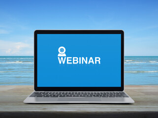 Webinar icon on modern laptop computer monitor screen on wooden table over tropical sea and blue sky with white clouds, Business seminar online concept