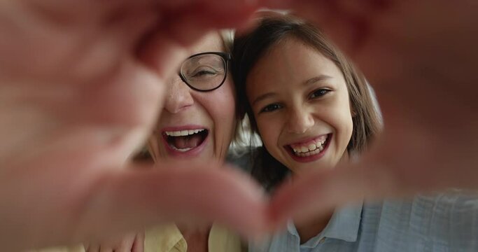 Close up happy faces of mature grandmother and pretty girl grandchild showing symbol of heart, laughing, look through fingers at camera. Sincere feelings, family ties and binding, unconditional love