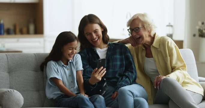 Three generational women, loving multigenerational family spend pastime use new mobile application on smart phone, laughing while watch funny on-line content enjoy weekend leisure on internet at home