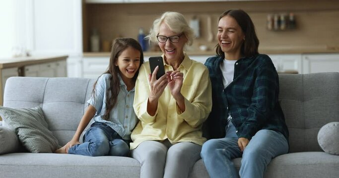 Happy mature 60s woman her daughter and grandchild use new mobile application, laugh on videos or funny filters use cellphone camera. Multigenerational family spend pastime with wireless modern tech
