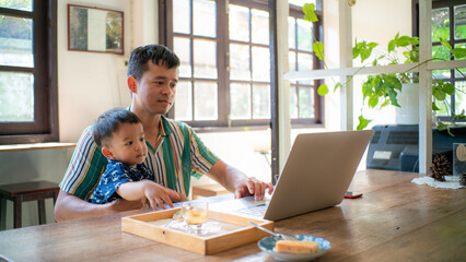 Asian daddy online working on laptop hold with toddler boy sitting in coffee shop
