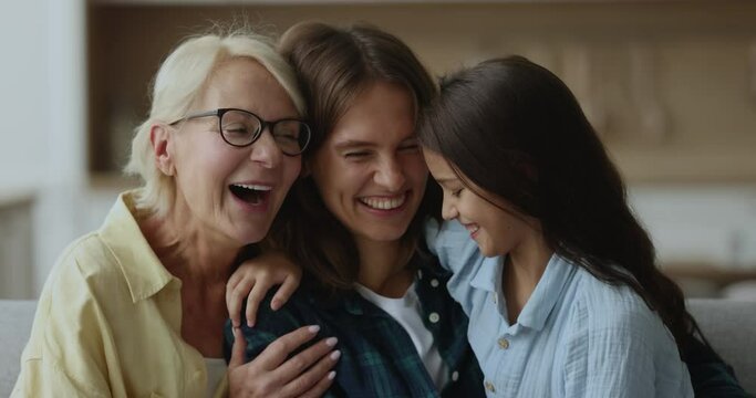 Laughing multi generational women cuddling, tickling each other, having fun seated on sofa at home, close up. Playful retired 60s grandma spend carefree leisure with grandkid and daughter. Family ties