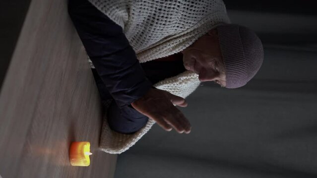 Old man dressed in a warm winter jacket sits at home at a table and warms his hands from a burning candle. Concept of no heating in winter at home.