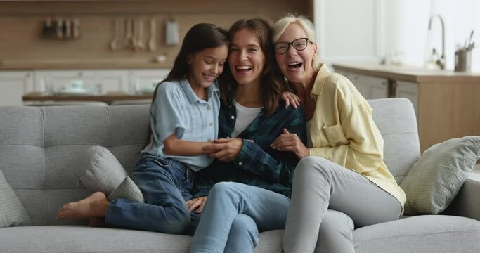 Three multigenerational women, happy family tickling each other, laughing seated on sofa smile look at camera. Older and younger relatives female enjoy playtime, spend playful leisure together at home