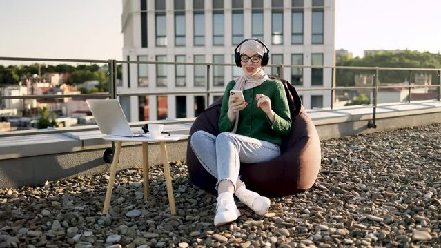 Emotional muslim lady in hijab singing along musical phrases in headphones while resting in pouf chair on terrace. Excited young woman rocking out to energetic tunes of favourite melodies outdoors.
