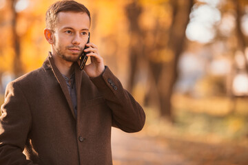 an attractive man in warm clothes walks in the autumn park and uses a mobile phone to talk.