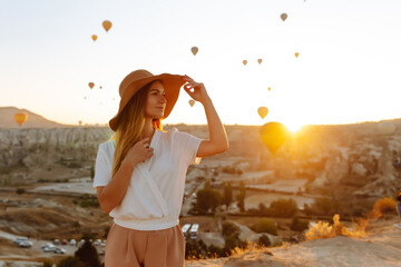 Happy woman stands on the mountain with flying air balloons on the background. Famous tourist...