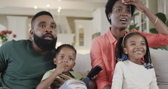 Excited african american parents with son and daughter watching sport on tv at home, slow motion