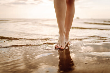 Close- up leg of young woman walking along wave of sea water and sand on the summer beach. Sexy lean and tanned legs. Summer holidays.
