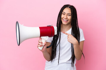 Teenager girl with braids over isolated pink background holding a megaphone and pointing side