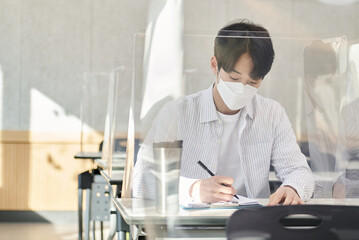A young male Asian college student is learning or listening to a lesson while her face is outside a protective shield on the desk in a university classroom in Korea during the pandemic.