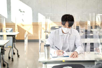 A young male Asian college student is learning or listening to a lesson while her face is outside a protective shield on the desk in a university classroom in Korea during the pandemic.