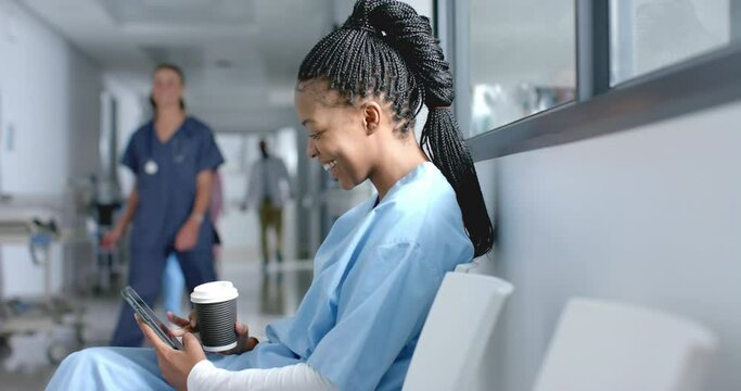 Happy african american female doctor using smartphone, drinking coffee in corridor, slow motion
