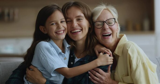 Close up of cheerful older 60s woman her young adult 35s daughter and pre-teen 10s girl, cuddling seated on couch smile look at camera, spend carefree happy weekend together at home, enjoy family ties