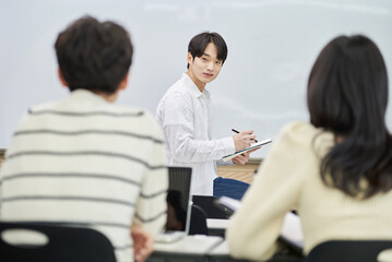 An Asian young man is standing in front of a lecture hall at a university in South Korea, giving a presentation or lecture. In front of him are male and female students. 