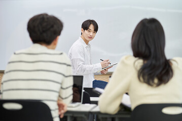 Fototapeta premium An Asian young man is standing in front of a lecture hall at a university in South Korea, giving a presentation or lecture. In front of him are male and female students. 