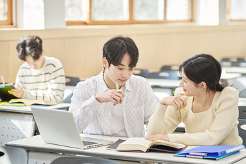 Fototapeta premium In a higher education classroom in South Korea, young university students are listening to a lecture, studying, and talking. A woman and a man are in the background