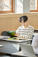 In a higher education classroom in South Korea, young university students are listening to a lecture, studying, and talking. A woman and a man are in the background