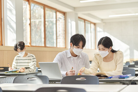In a higher education classroom in South Korea, young university students are listening to a lecture, studying, and talking. A woman and a man are in the background