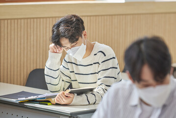 In a higher education classroom in South Korea, young university students wearing masks are listening to a lecture, studying, and talking. A woman and a man are in the background