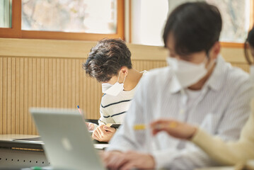 Fototapeta premium In a higher education classroom in South Korea, young university students wearing masks are listening to a lecture, studying, and talking. A woman and a man are in the background