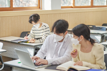 Obraz premium In a higher education classroom in South Korea, young university students wearing masks are listening to a lecture, studying, and talking. A woman and a man are in the background