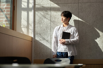 Picture of a young man who is a college student standing against a wall in a lecture hall at a university in South Korea. The room is bright