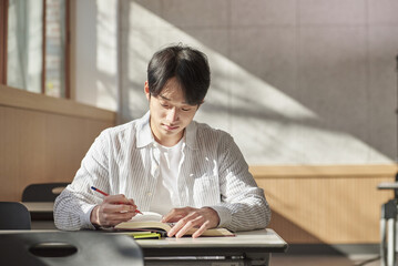 A young college student is sitting at a desk in a university classroom in South Korea, Asia. He is either reading a book or looking out of the window