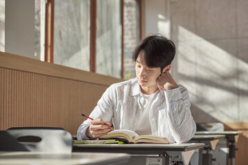 A young college student is sitting at a desk in a university classroom in South Korea, Asia. He is either reading a book or looking out of the window