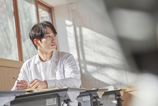 A young college student is sitting at a desk in a university classroom in South Korea, Asia. He is either reading a book or looking out of the window