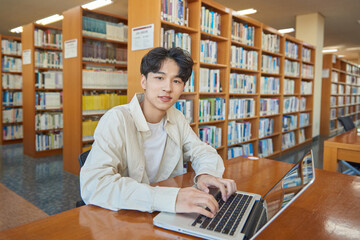 A male student sits at a desk in a university library in South Korea, Asia, looking at a laptop...