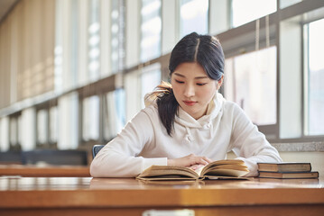 Asian Korean University Library Young female college student model sitting at a desk by a window with light coming in, reading a book, looking out the window
