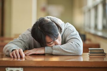 Fototapeta premium Asian Korean University Library Young male college student model sitting at a desk by a window with light coming in, reading a book, looking out the window or leaning down.