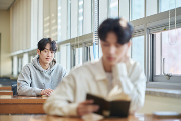 Three college students, both male and female, are sitting or standing at a desk in a library at a South Korean university in Asia, looking at laptops and books while having a conversation.