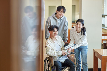 A young male model with a disability sitting in a wheelchair reading a book in a university library in Asia, with two young male and female college students helping him