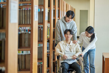 A young male model with a disability sitting in a wheelchair reading a book in a university library in Asia, with two young male and female college students helping him