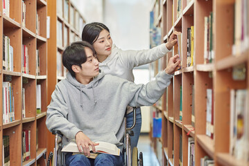 A young male college student model with a physical disability in a wheelchair looking for a book on a shelf in a university library in South Korea. A female college student model helping him