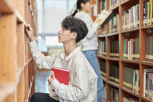 Two young male and female college students looking for and looking at books on the shelves of a university library in South Korea, Asia