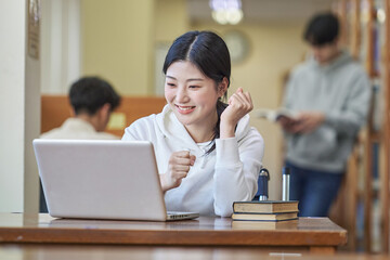 Young Asian Korean female model in library looking at laptop or book, lecture, assignment, discussion, male model in background, bookshelf in background