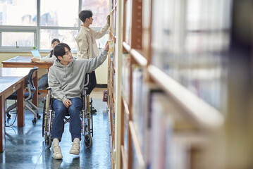 Young handicapped disabled male college student model in wheelchair looking for books in library bookshelf at university in Asia, helping male college student