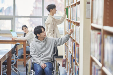 Young handicapped disabled male college student model in wheelchair looking for books in library...