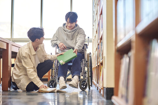 Young Handicapped Disabled Male College Student Model In Wheelchair Looking For Books In Library Bookshelf At University In Asia, Helping Male College Student