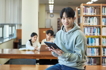 Young Asian Korean male model, man and woman model and looking out the window with tablet in the library on the background of bookshelf