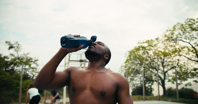 A man with Black skin and a bare torso drinks water on a basketball court and then licks her head to cool off and shouts his battle cry in front of his friends playing basketball