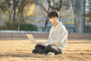 Young and attractive college student male model sitting on lawn at Asian Korean university in autumn, looking at laptop, listening lecture, relaxing or talking online.