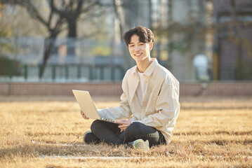 Young and attractive college student male model sitting on lawn at Asian Korean university in autumn, looking at laptop, listening lecture, relaxing or talking online.