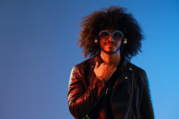 Portrait of a stylish man with curly hair with glasses and headphones on a blue background multinational, colored light, black leather jacket trend, modern concept.