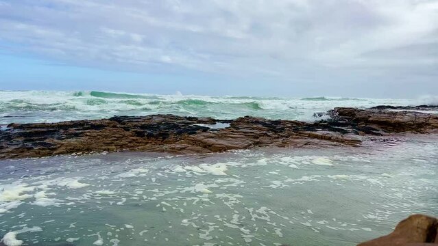 Slow motion of the foamy waves of the sea hitting the rocky shore under the cloudy sky