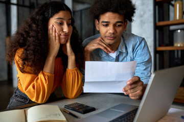 Upset worried shocked black female touching her face while looking at screen of laptop sitting next to her husband holding invoice letter or bill for electricity, reading attentively